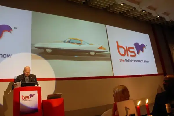 Speaker at a bis lectern in front of screens showing The British Invention Show