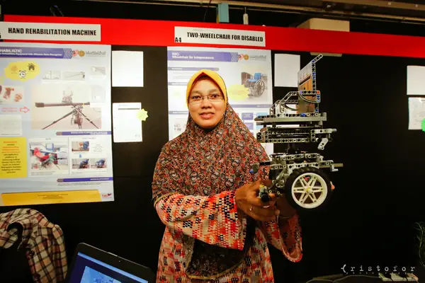 Exhibitor holding a model wheelchair beside posters for a hand rehabilitation machine and a two-wheelchair for the disabled